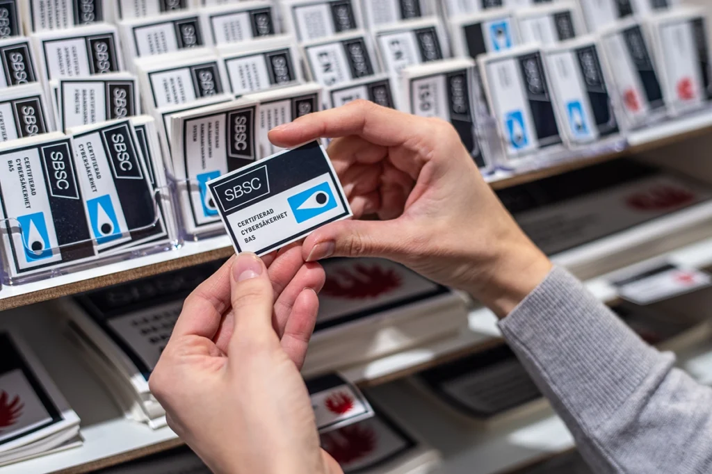Händer håller ett SBSC-certifieringsmärke för Cybersäkerhet BAS framför en display med certifieringsmärken.